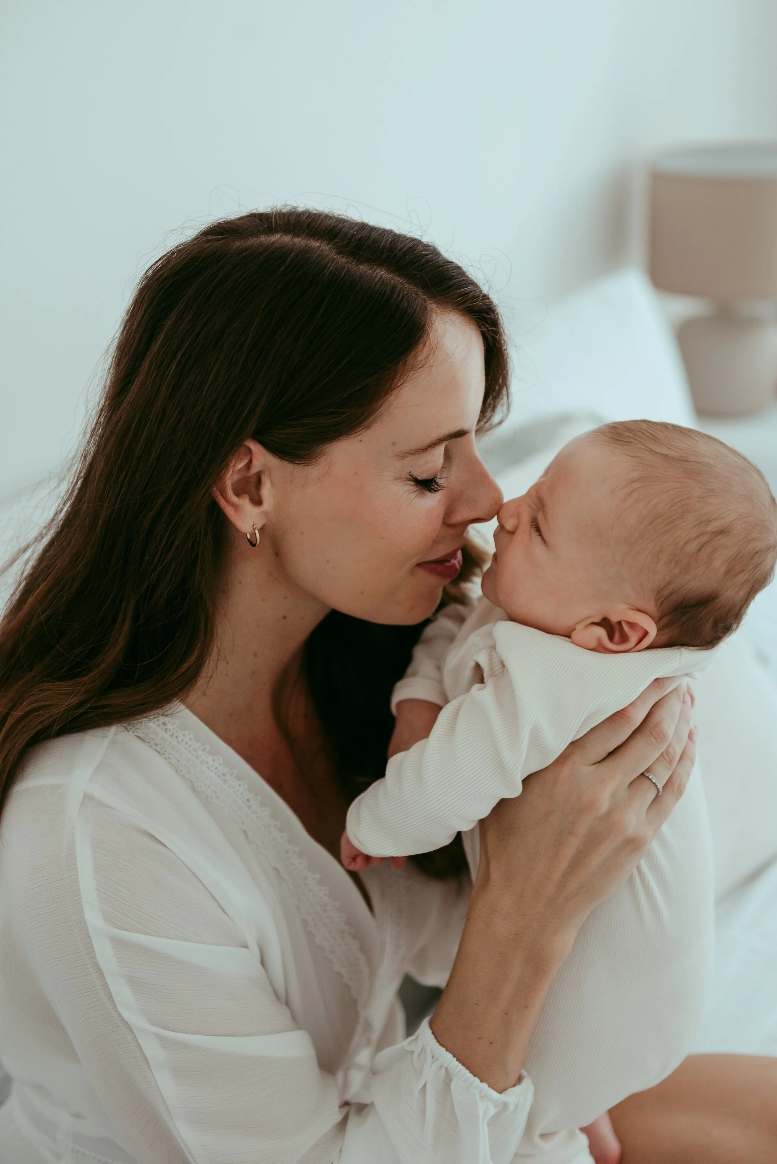 A woman with dark hair and earrings holds a baby close to her face, both gently touching noses in a white room.