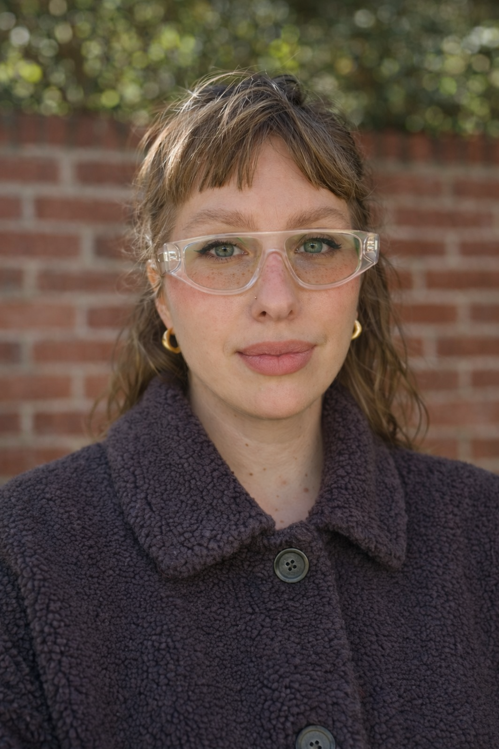A woman with light brown curly hair, wearing clear safety glasses, gold hoop earrings, and a dark purple fuzzy coat, standing outdoors in front of a brick wall with greenery in the background.