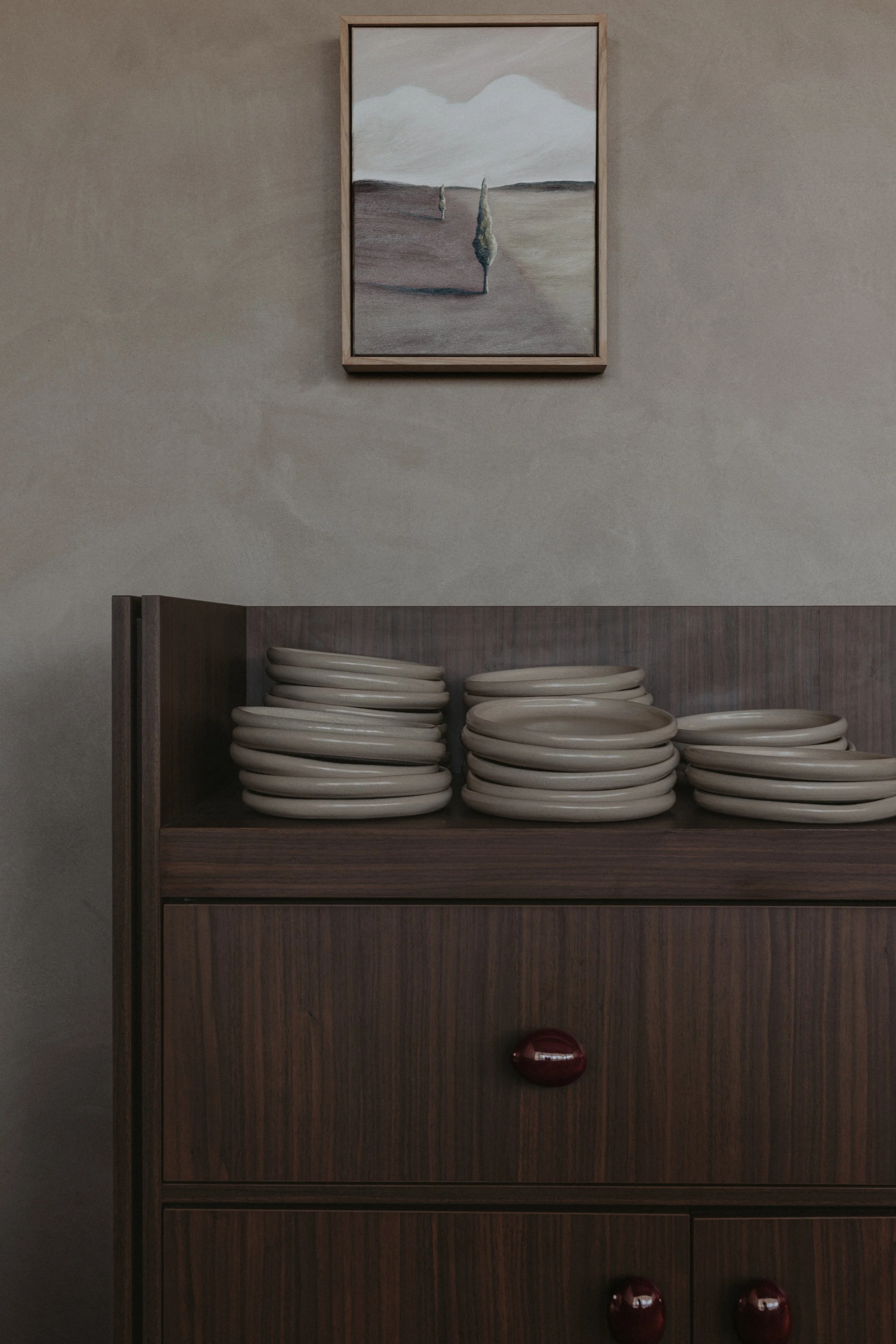 A wooden cabinet with stacks of beige plates on top and a framed landscape painting with cypress trees on a neutral wall above.
