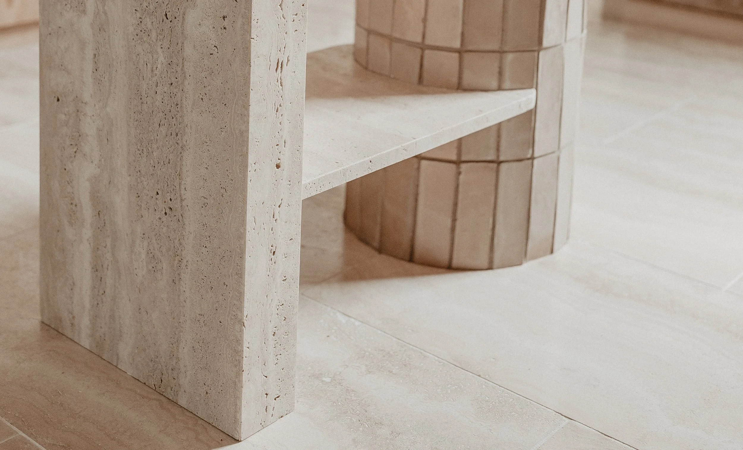 Close-up of a natural travertine stone bench with a vertical edge, and a rounded side with a handmade tiled pattern, on a matching travertine tiled floor.