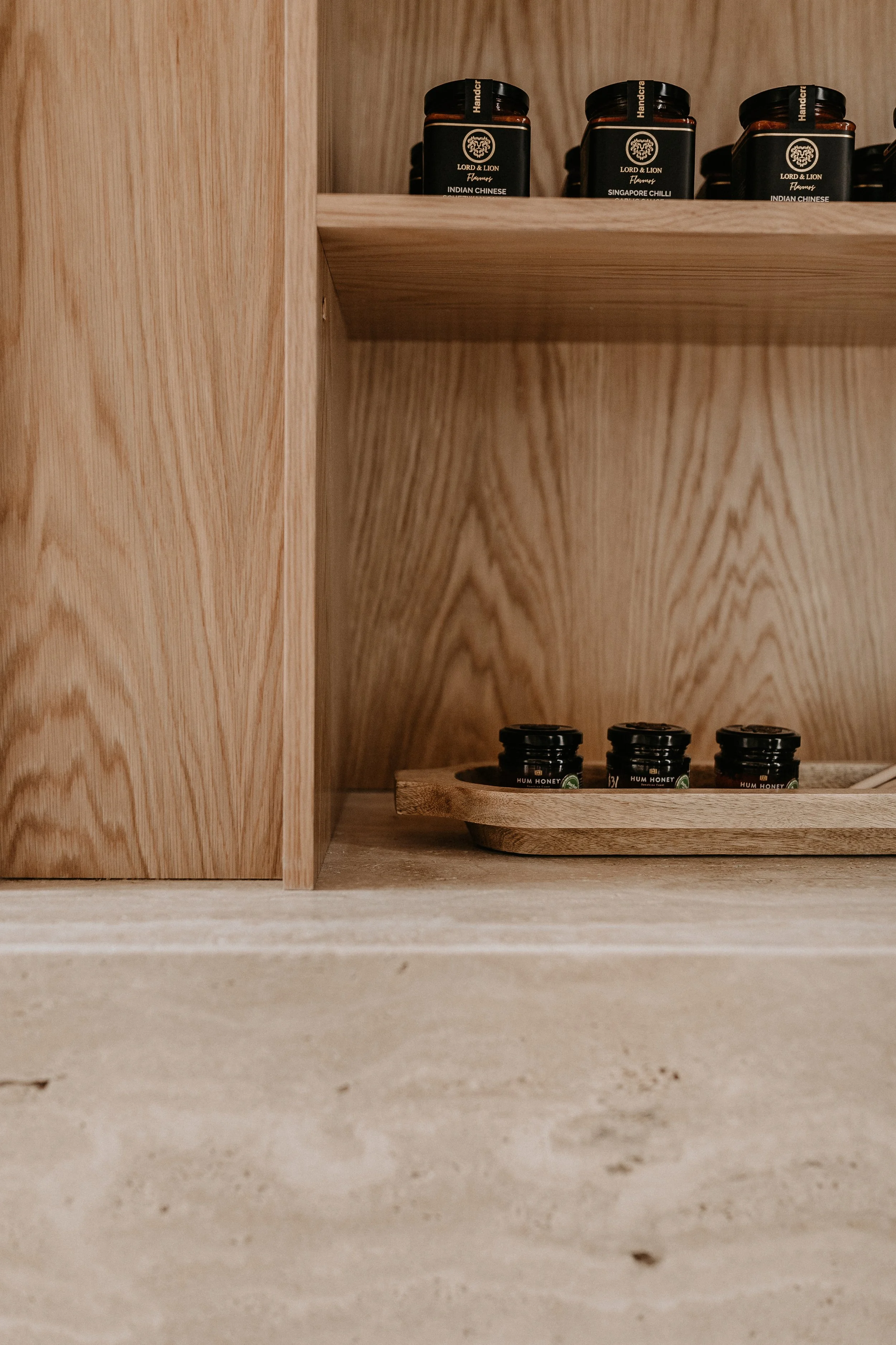 Close-up of a wooden kitchen shelf with black jars labeled 'Indian Chinese' and 'Singapore Chilli,' and small containers labeled 'HUM HONEY' on a wooden tray.