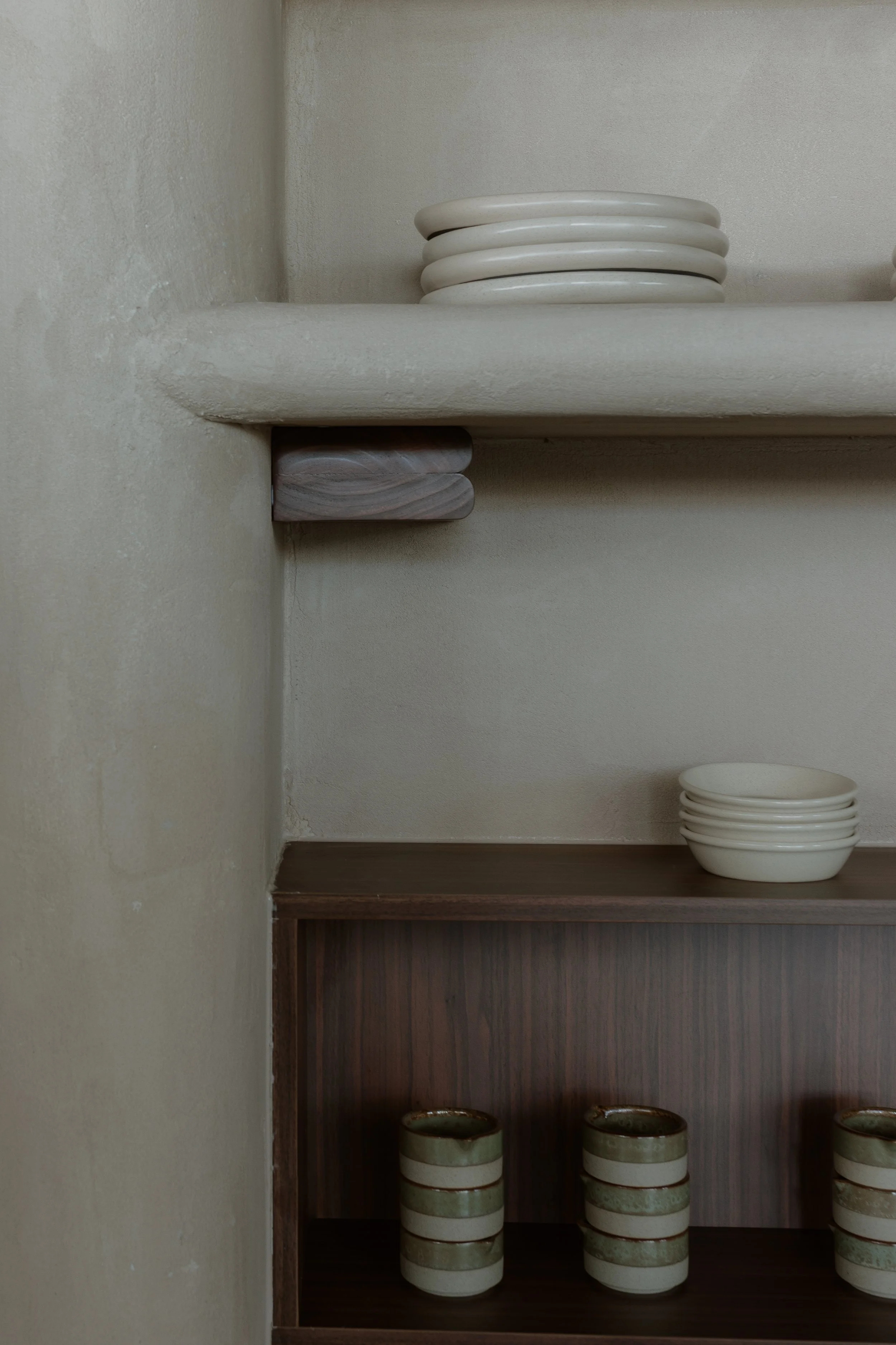 Kitchen shelf with stacked white plates on top, a stack of three small bowls on the middle shelf, and two groups of three smaller green and brown cups on the bottom shelf.
