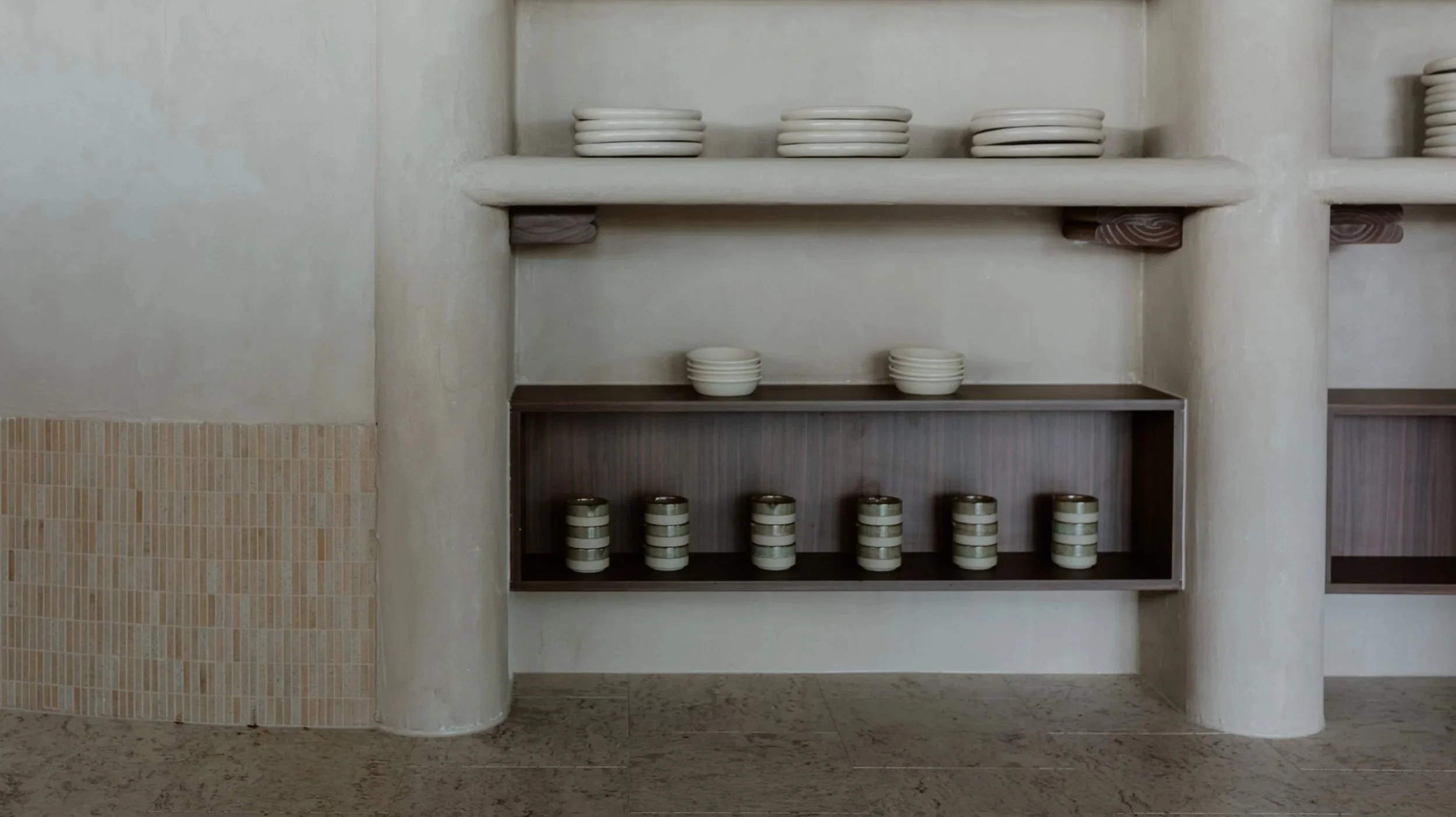 Ceramic plates and bowls on shelves and at the bottom, decorated containers with horizontal stripes, set in a minimalist, neutral-toned room.