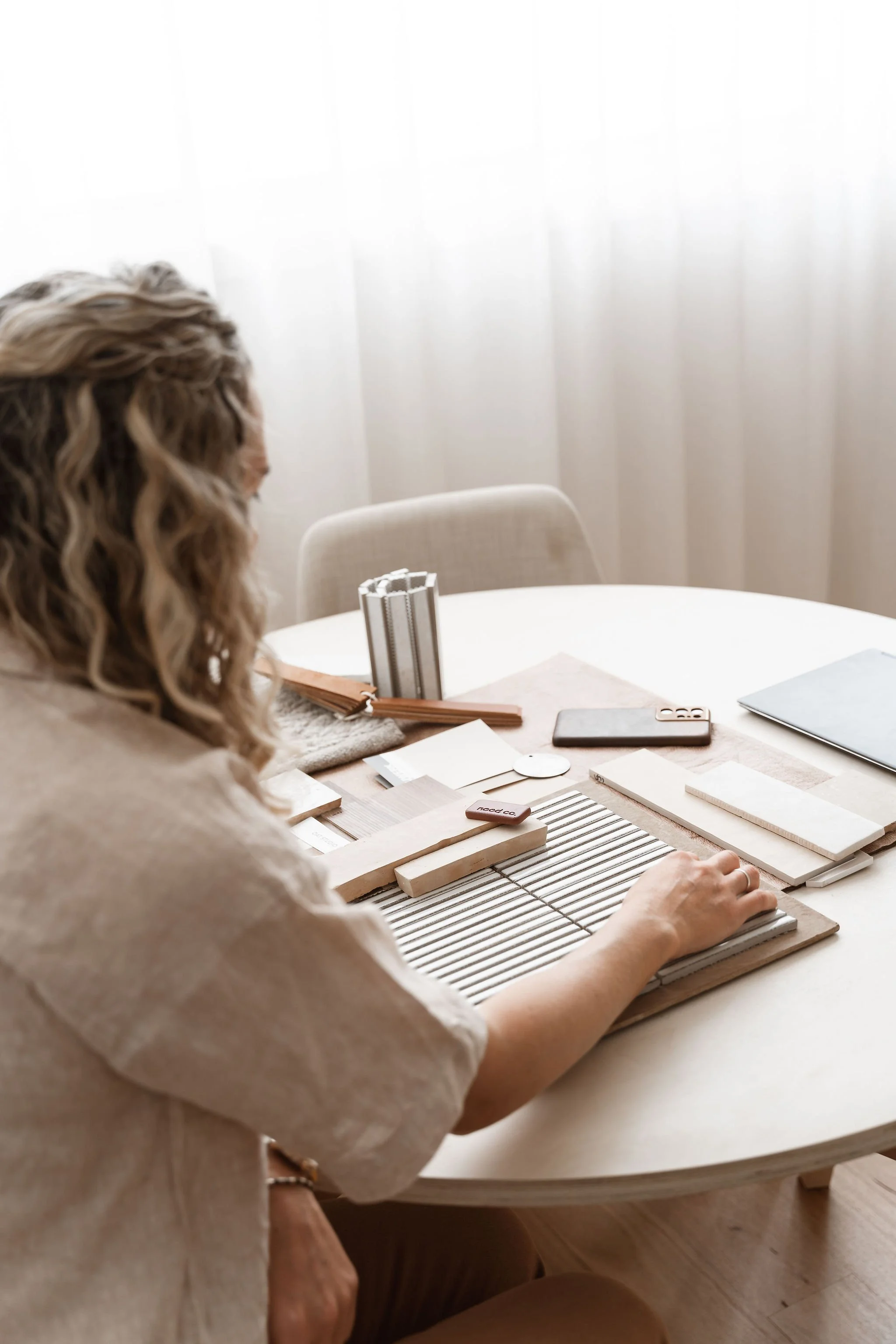A woman with curly hair sitting at a round table with various design and color samples, a phone, and a laptop, in a bright room with sheer curtains.