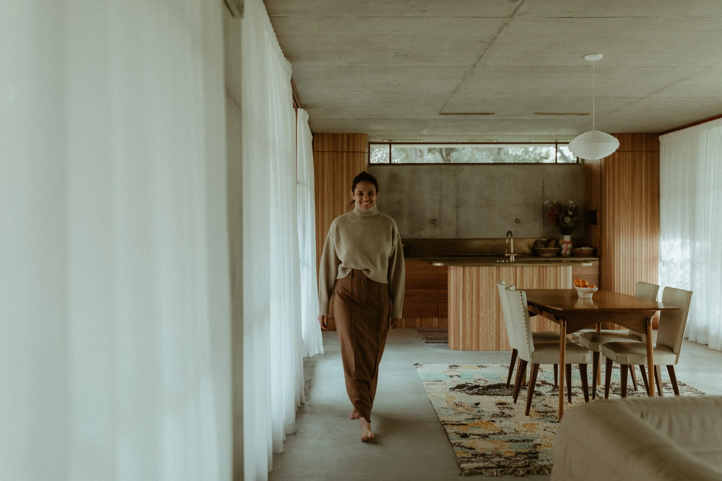 A woman walking barefoot in a cozy, modern kitchen and dining area with natural light, beige curtains, a wooden dining table with six chairs, and a kitchen island with a sink, countertop, and a flower vase in the background.