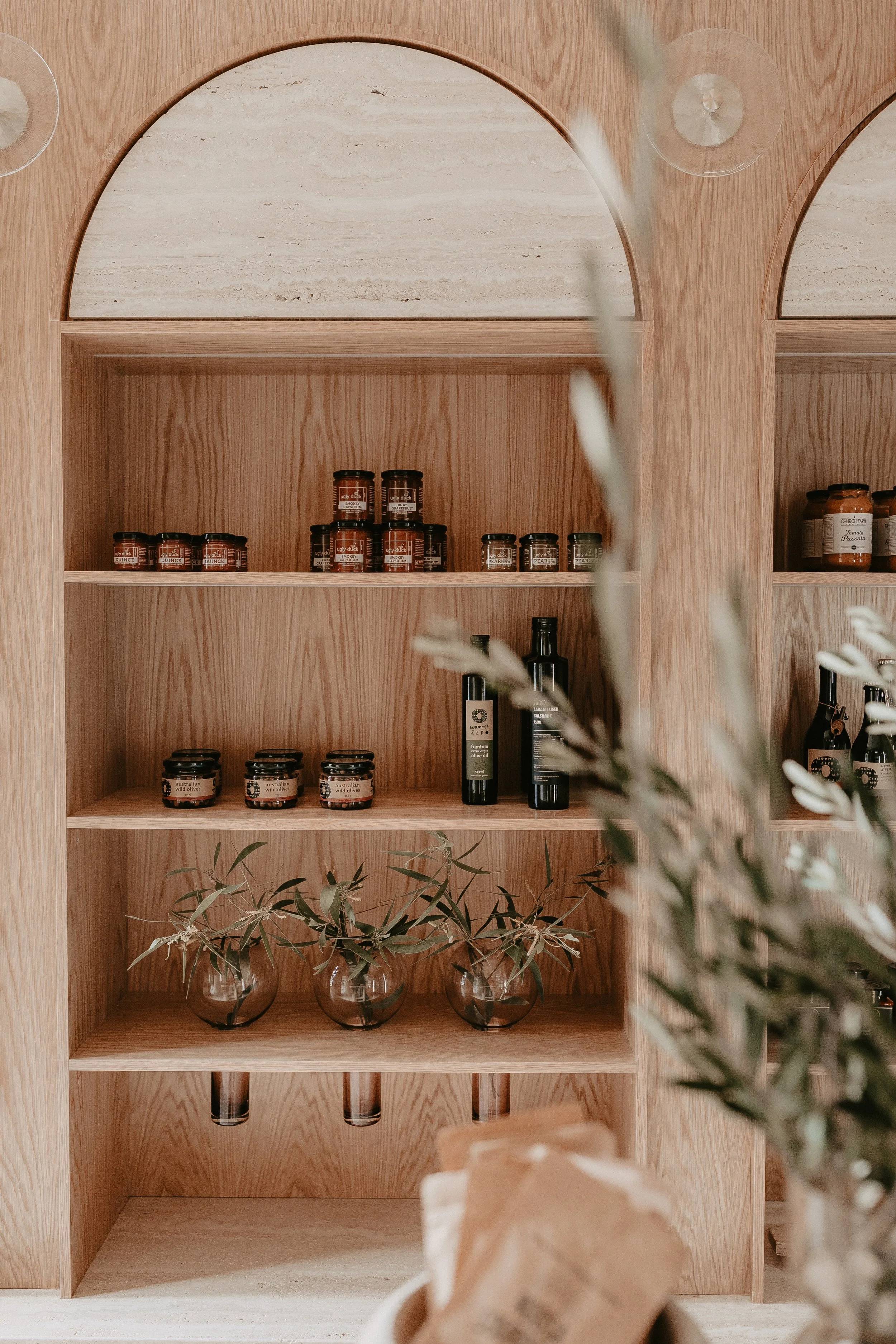 Wooden shelf with jars of food items, a few bottles, clear glass vases with green leafy branches, and a blurred plant in the foreground.