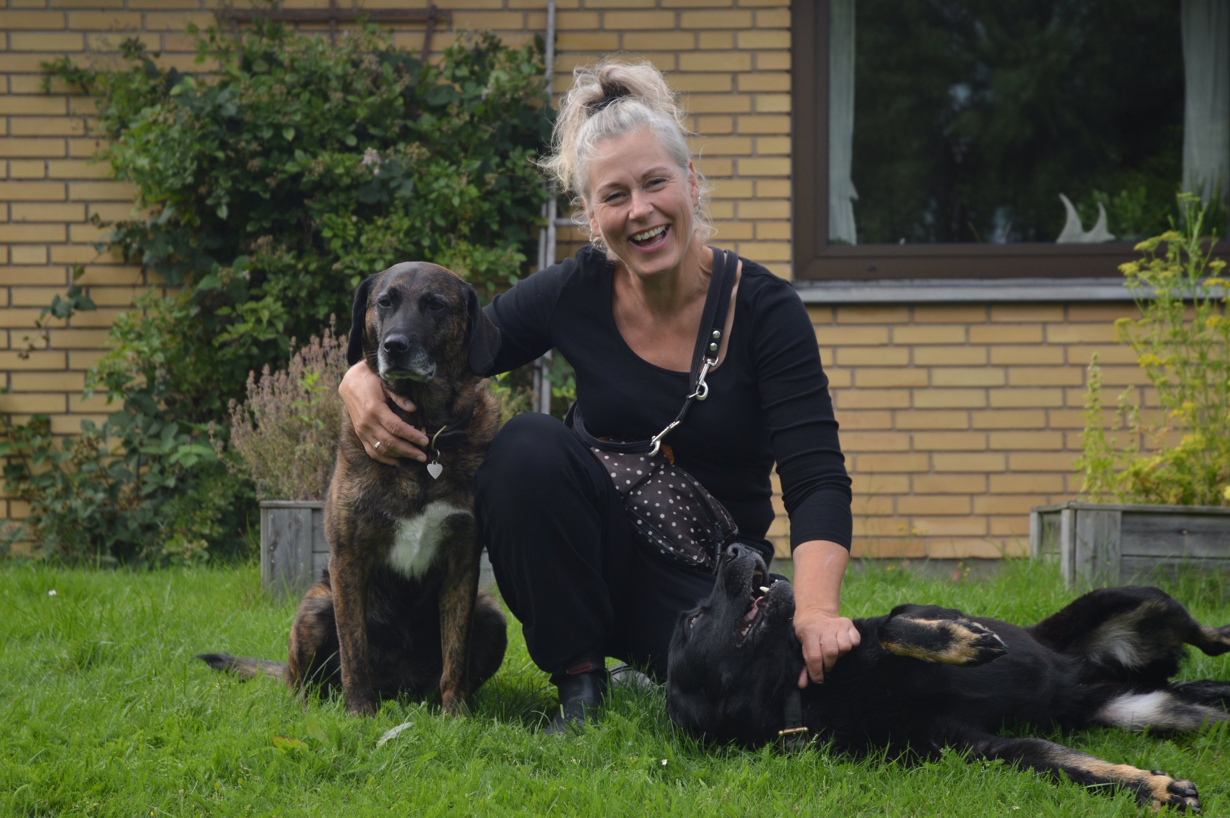En glad kvinde leger med to hunde i en have foran en murstensvæg.