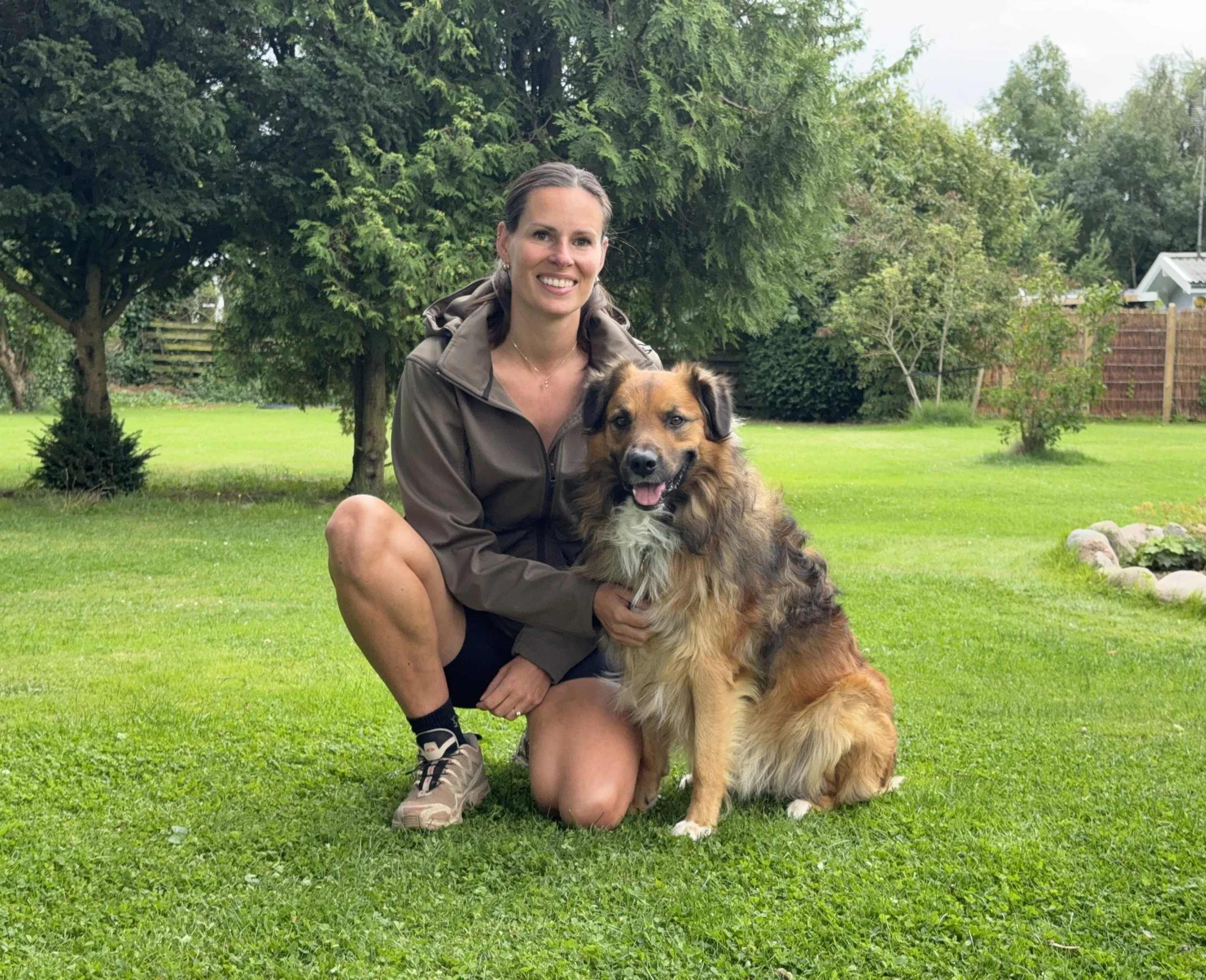 En smilende kvinde i udendørs omgivelser med grønt græs og træer, som kneler ved siden af en blandet race hund med lang pels.