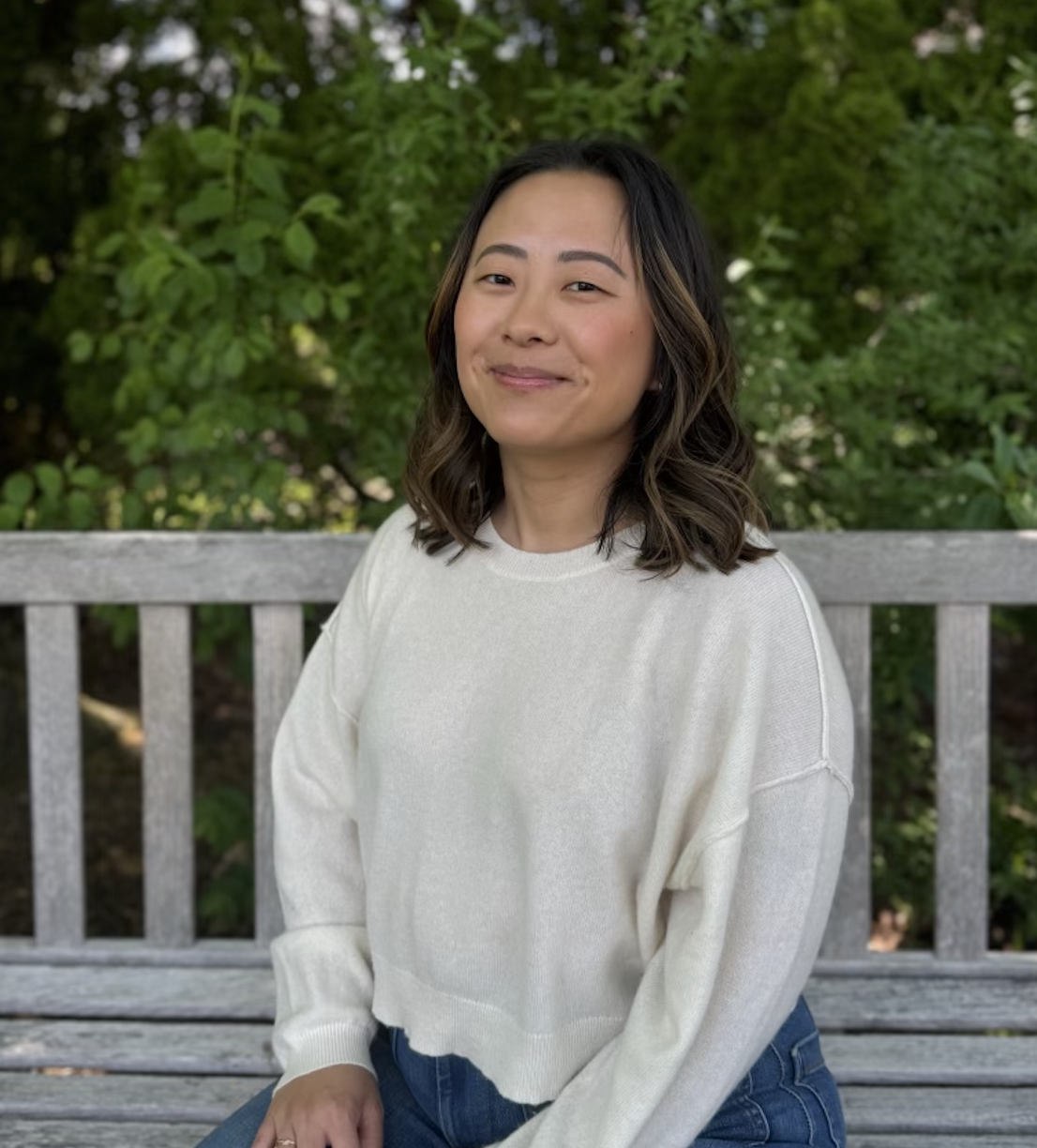 A woman with wavy dark hair and a white sweater sitting on a wooden bench outdoors with green foliage in the background.