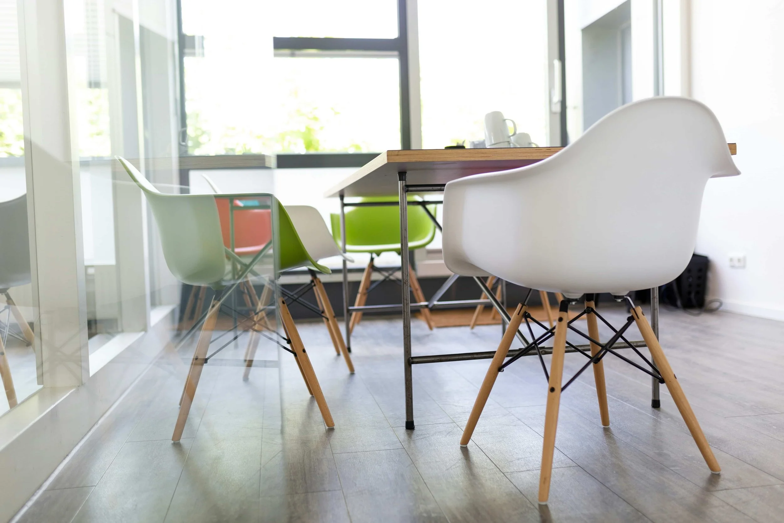 Modern conference room with a large wooden table and colorful plastic chairs, with large windows allowing natural light to fill the room.