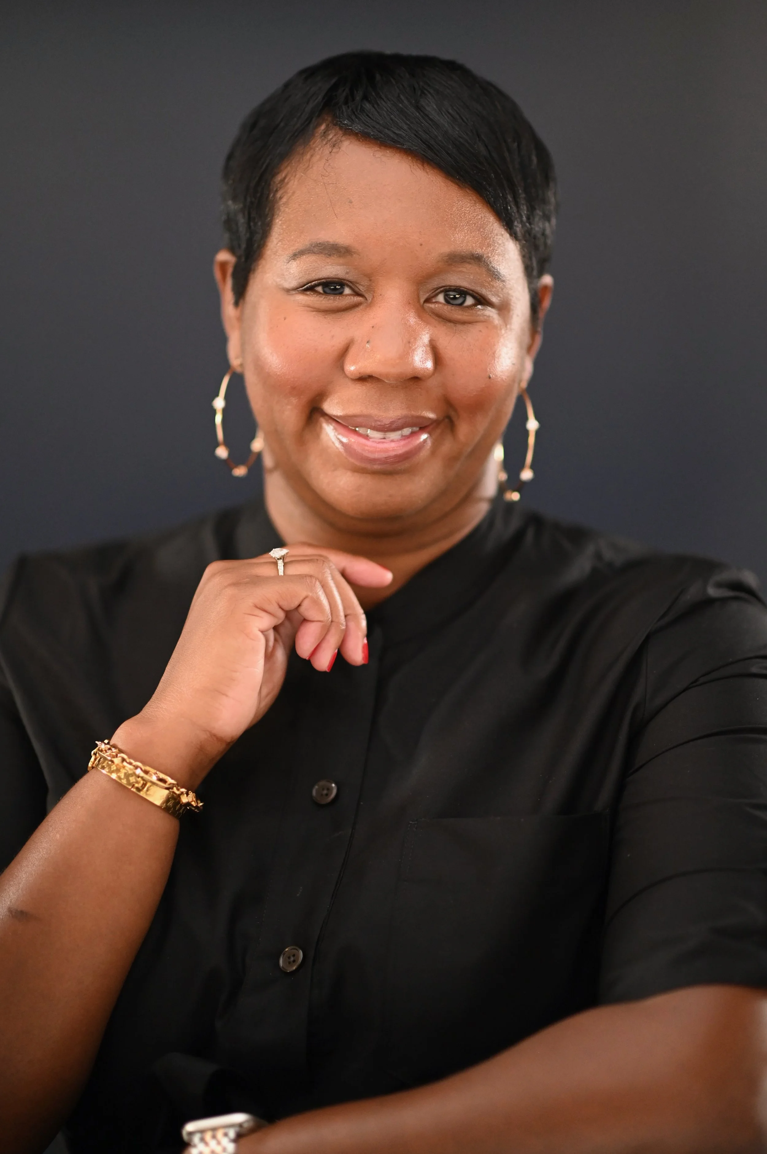 A woman with short black hair and gold jewelry, including hoop earrings, a bracelet, and a ring, wearing a black button-up shirt, posing against a dark background.
