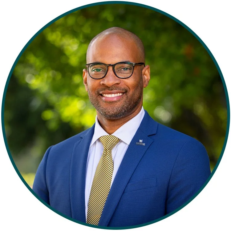 A professional man with a bald head, glasses, and a beard, wearing a dark blue suit, a white dress shirt, and a yellow striped tie, smiling outdoors with blurred green trees in the background.