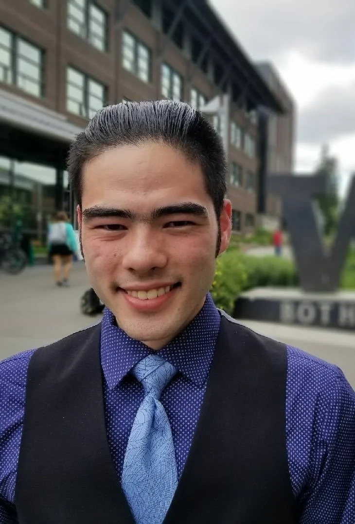 A young man with slicked-back hair smiling at the camera, wearing a blue shirt, a light blue tie, and a black vest. He is standing outside a modern brick building with large windows, with people walking in the background.