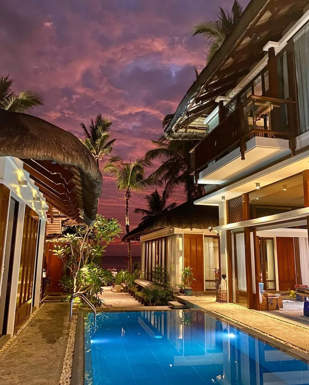 A tropical resort with a swimming pool, surrounded by lush plants and thatched-roof structures, under a colorful sunset sky with pink and purple clouds and palm trees.  Harlan Beach Villa Boracay