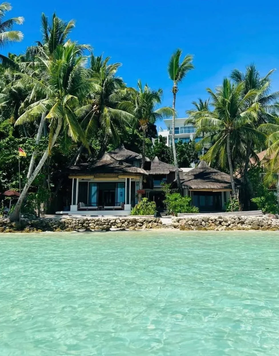 A tropical beach scene with clear turquoise water in the foreground, and a thatched-roof bungalow surrounded by palm trees against a bright blue sky. Harlan Beach Villa Boracay
