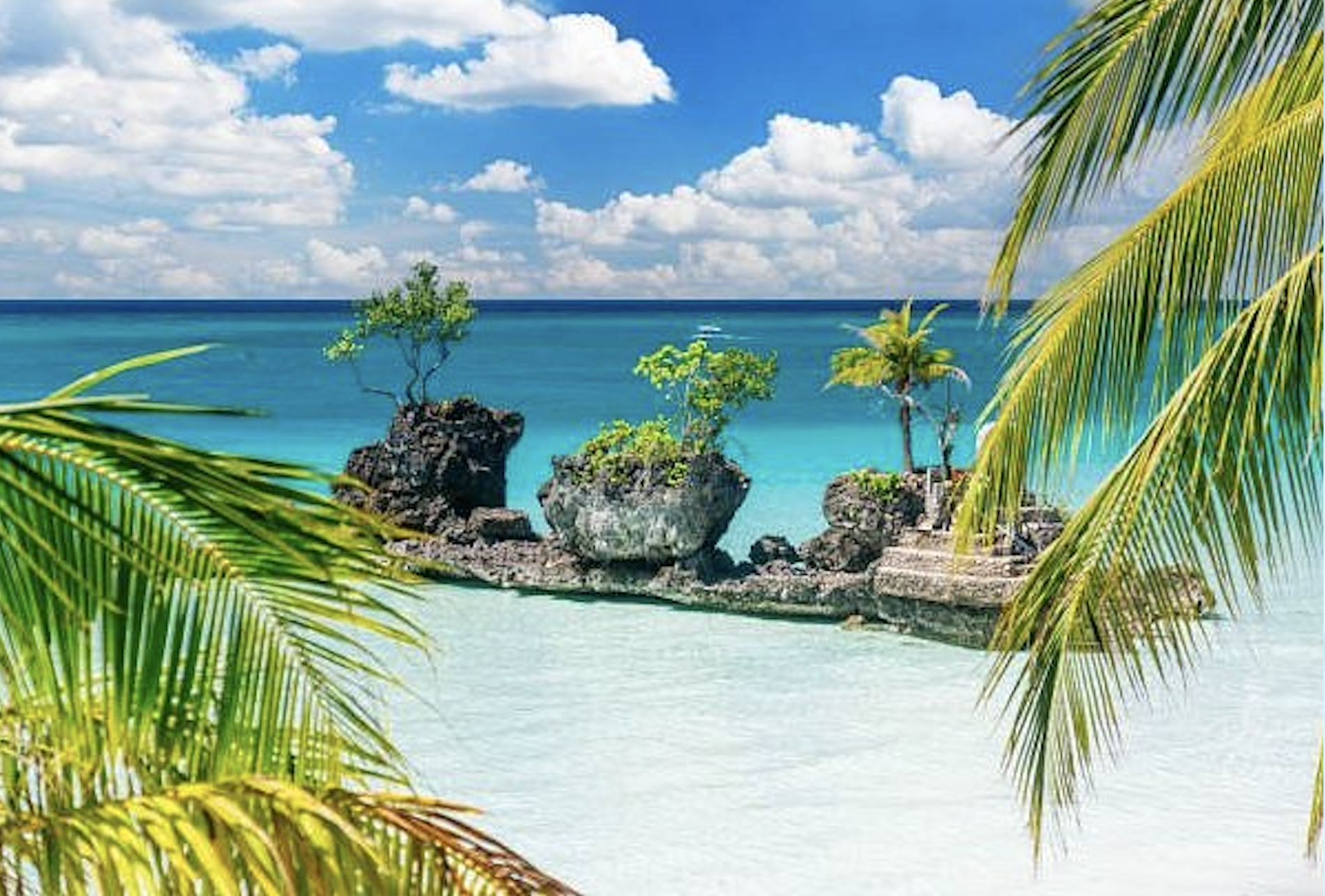 Tropical beach with white sand, turquoise water, rocky formations with small trees, and palm fronds in the foreground. Harlan Beach Villa Boracay