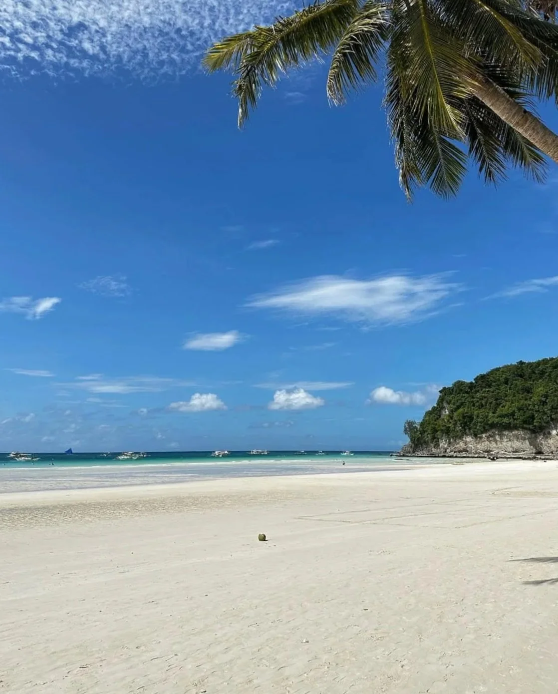 A scenic tropical beach with white sand, clear blue water, and a partly cloudy sky. Palm trees and a distant green hill are visible. Harlan Beach Villa Boracay