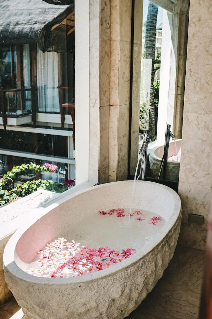 Stone bathtub filled with water and floating pink and white flowers, located near a large window with a view of a balcony and tropical plants. Harlan Beach Villa Boracay