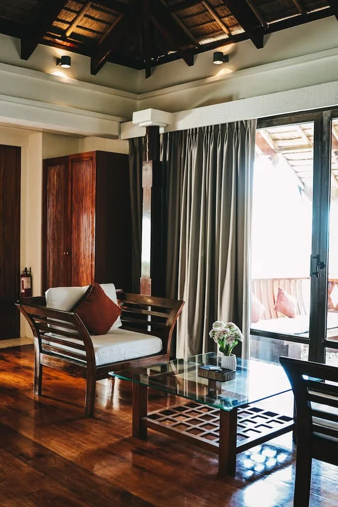 Interior of a living room with wooden furniture, beige curtains, a glass coffee table with a flower vase, and a sliding glass door leading to a balcony. Harlan Beach Villa Boracay