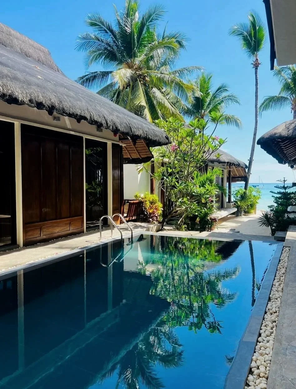 Resort with thatched-roof huts, a swimming pool, palm trees, and clear blue sky by the beach. Harlan Beach Villa Boracay