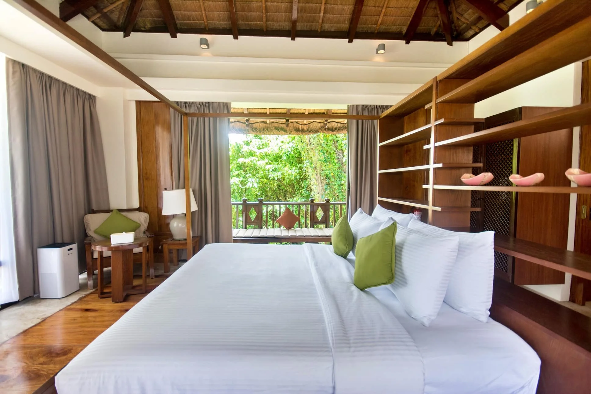 A bright hotel room with a large bed and white linens, green pillows, and a wooden headboard with open shelves. The room has a balcony with greenery outside, beige curtains, and wooden furniture including a side table with a lamp and a bench with a c