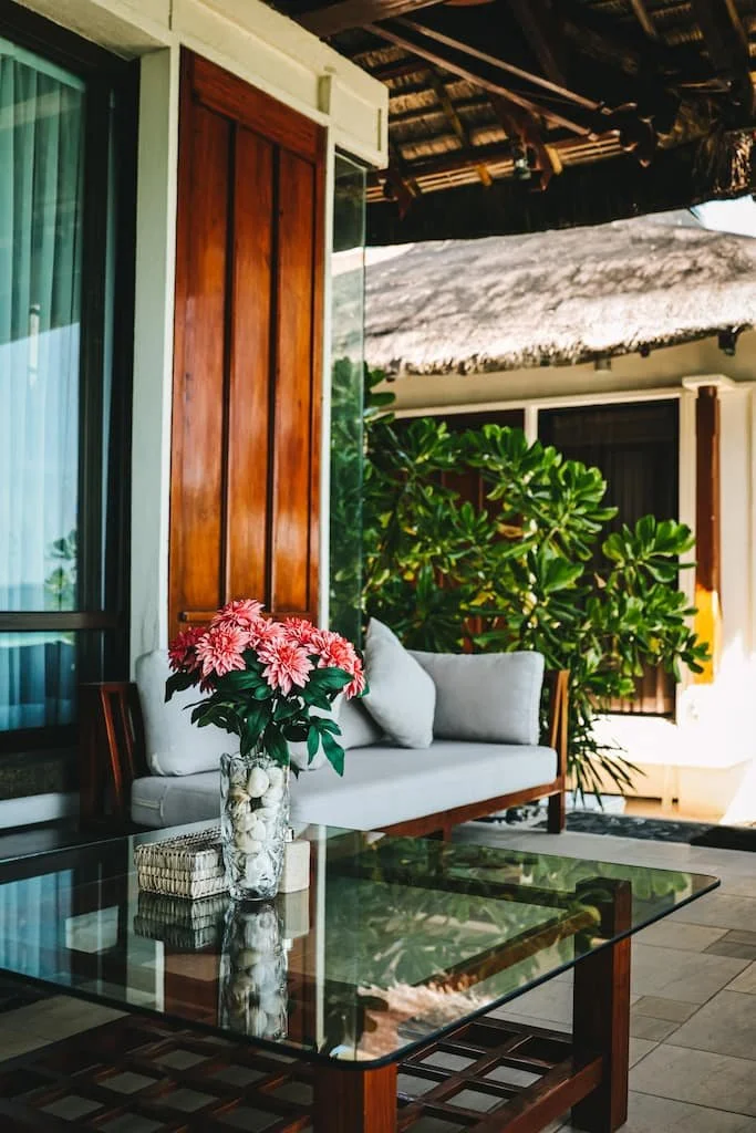 Outdoor patio with a glass-top table, a white cushioned bench with pillows, a vase of pink flowers, green plants, wooden walls, and a thatched roof overhead.  Harlan Beach Villa Boracay