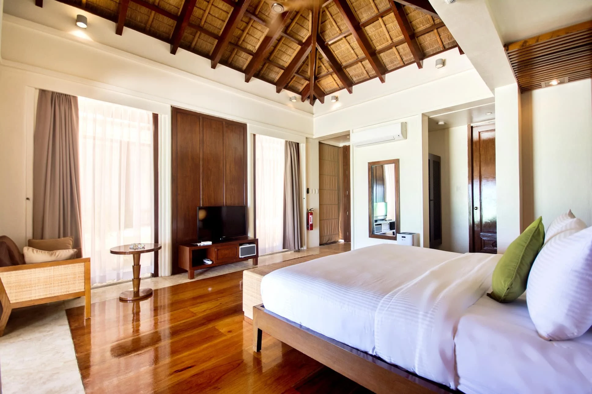 Hotel room with a large bed, wooden furniture, a flat-screen TV, a mirror, and large windows with curtains, featuring a thatched ceiling and wooden accents. Harlan Beach Villa Boracay