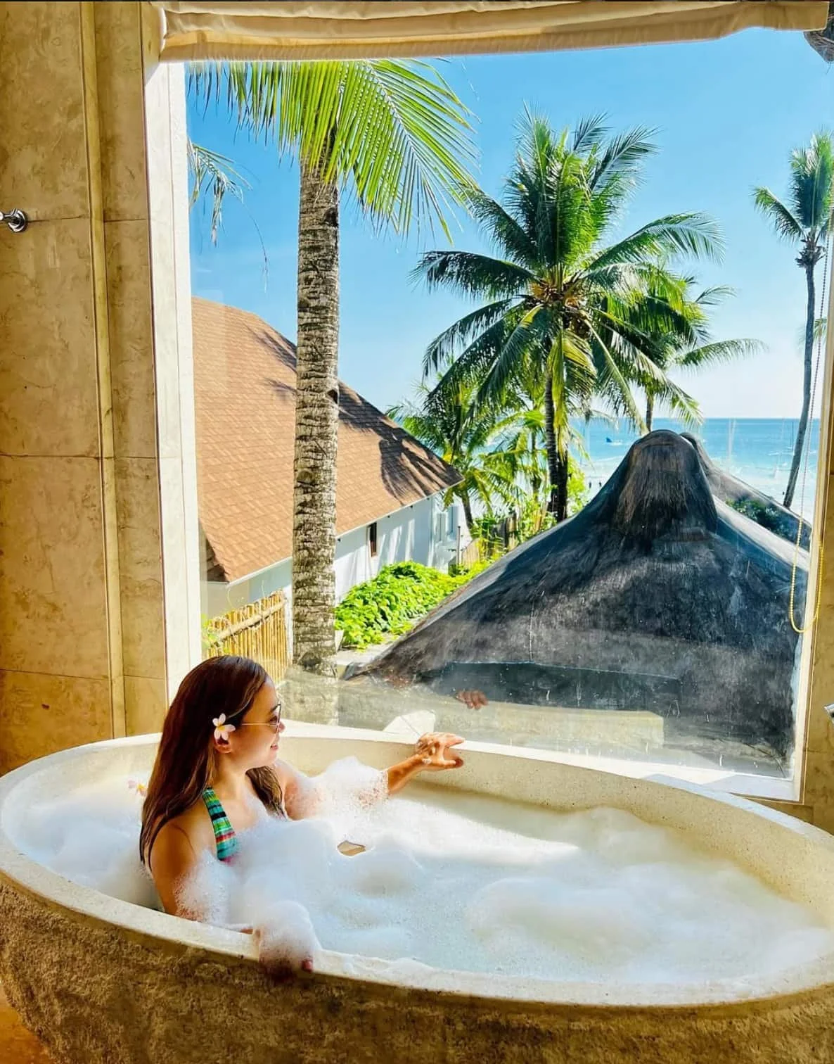 A woman relaxing in a bubble bath with a view of palm trees, a thatched roof, and the ocean through a large window. Harlan Beach Villa Boracay