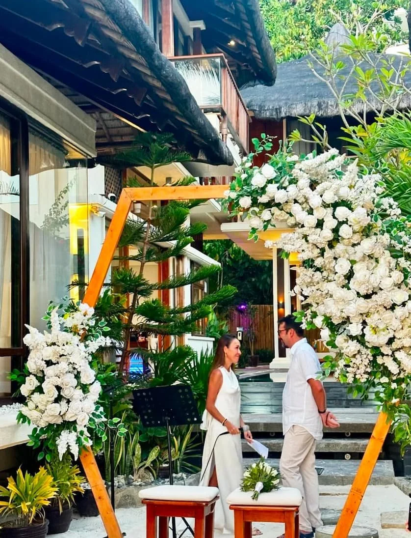 A couple has a conversation at an outdoor wedding altar decorated with white flowers and green foliage. The setting appears tropical or coastal with lush plants and a wooden house in the background.