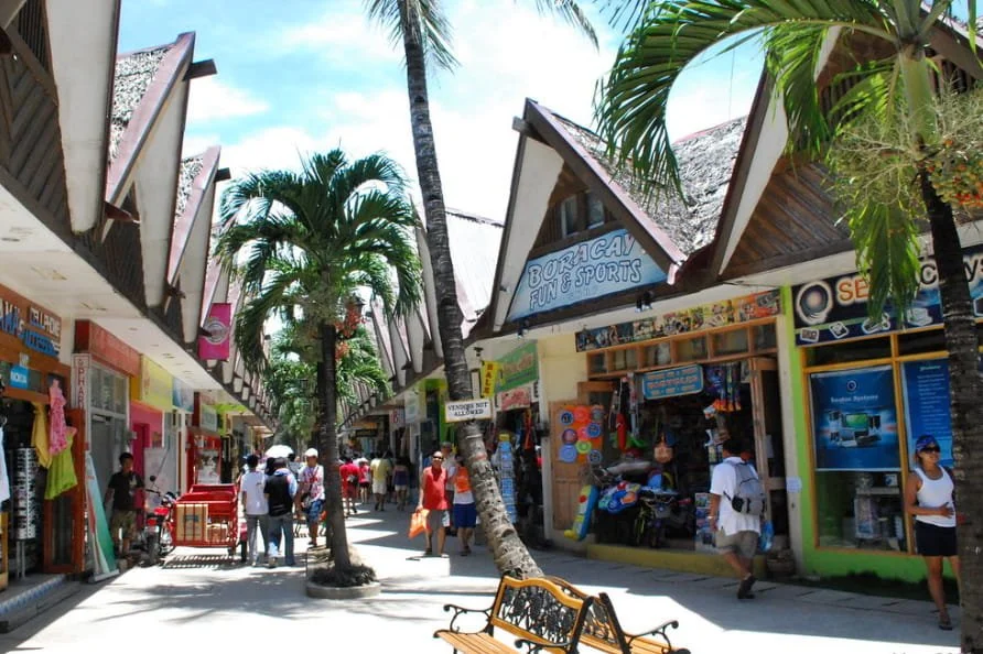 Street scene in a tropical location with shops on either side and palm trees in the middle. People are walking, shopping, and sightseeing.
