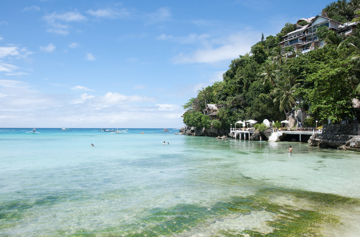 Tropical beach with clear turquoise water, palm trees, and hillside resort buildings with balconies and umbrellas. Harlan Beach Villa Boracay