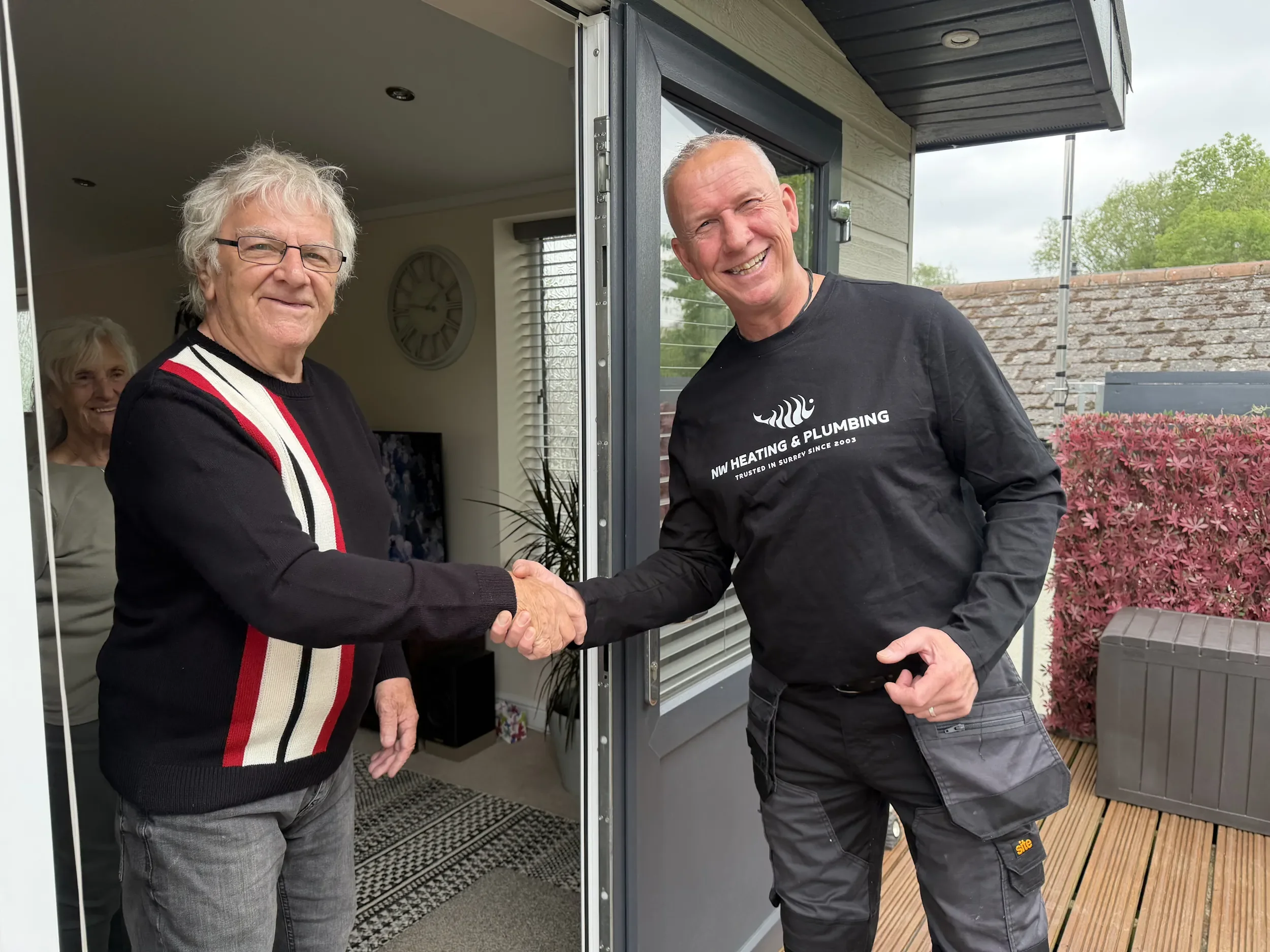 Two men shaking hands at the entrance of a house, smiling. One man wears glasses and a black sweater with red, white, and beige stripes. The other man is wearing a black T-shirt with white text and gray pants. Inside, a woman with blonde hair is visible, and a clock hangs on the wall.