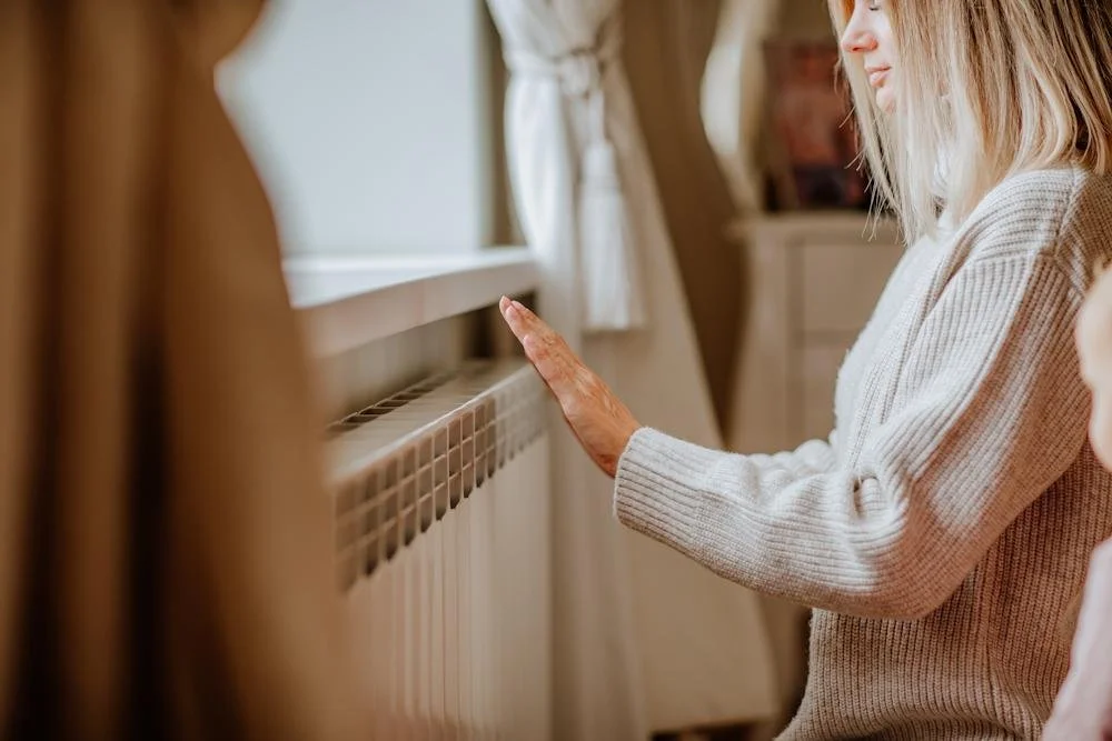 A woman softly touching a radiator with her hand, indoors near a window with white curtains.