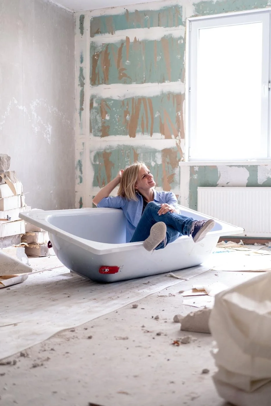 A woman sitting relaxed in an empty bathtub in a room under renovation, with unfinished walls and construction debris on the floor, bright natural light coming through a window