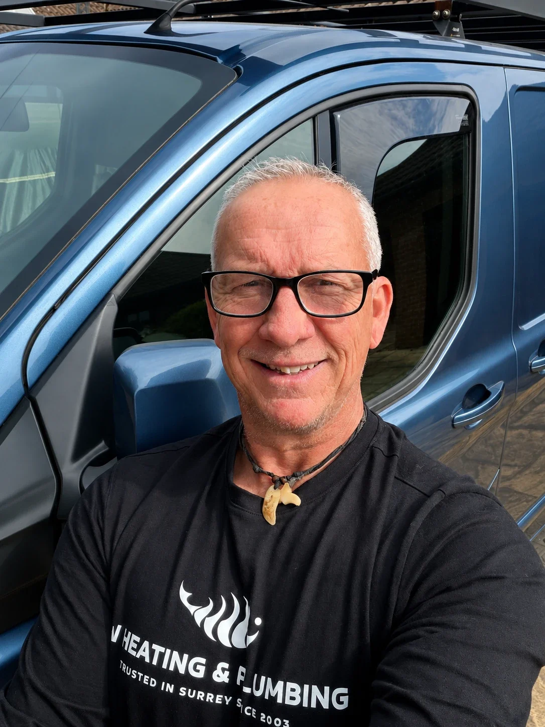 A smiling man with glasses, wearing a black t-shirt and a necklace with a claw pendant, taking a selfie in front of a blue vehicle.