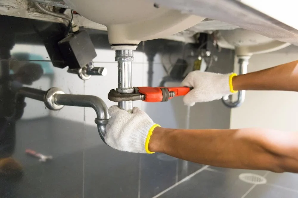 Person wearing white gloves using a pipe wrench to tighten a pipe connection under a sink.