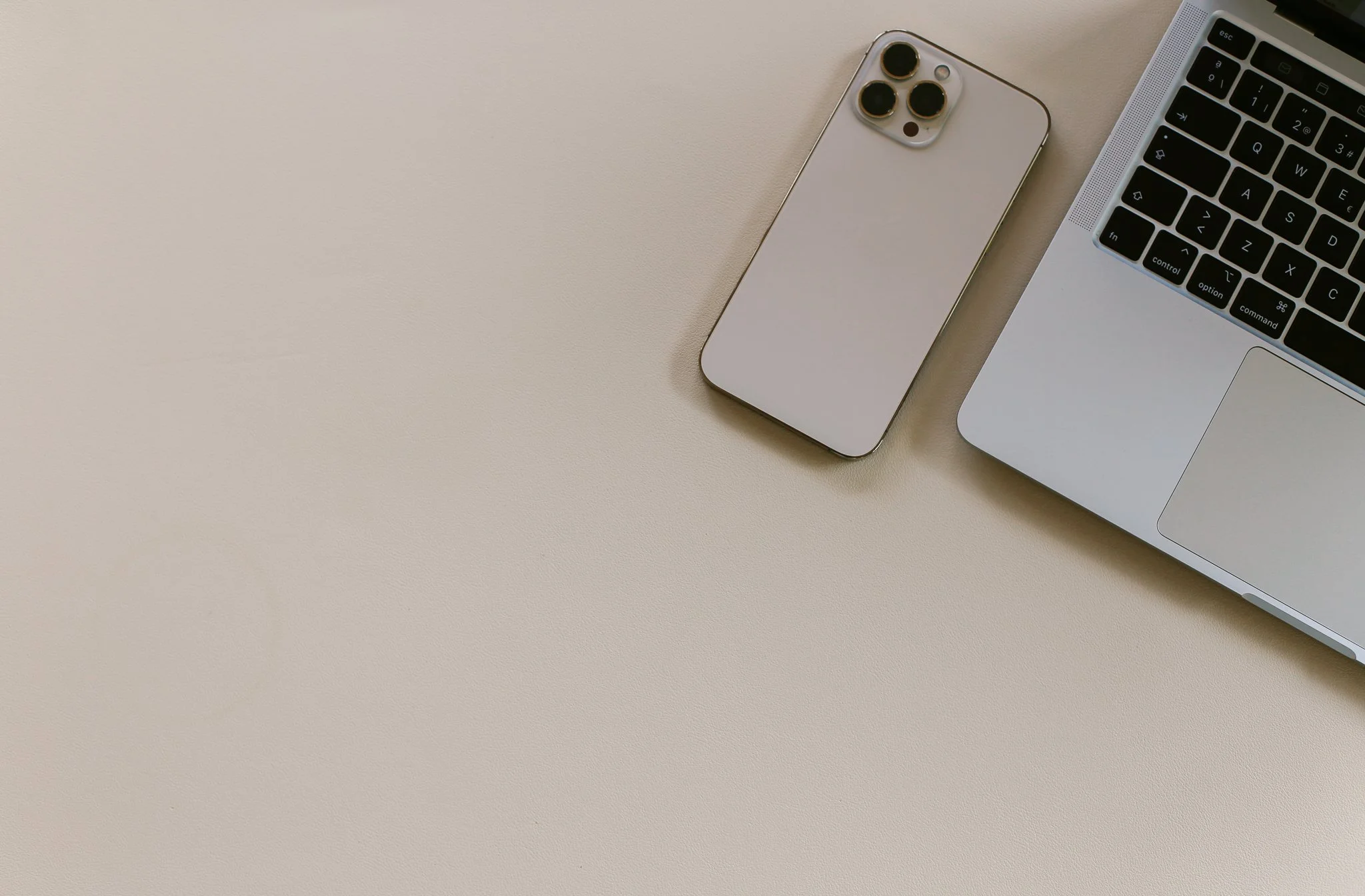 An open silver laptop with a black keyboard, a white smartphone with a triple camera, all on a beige surface.
