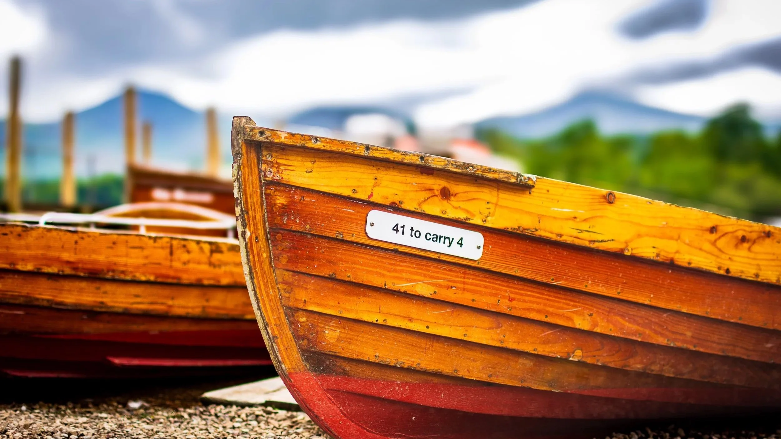 Close-up of a vintage wooden boat's bow with a sign that reads "41 to carry 4" on the side, set outdoors with other boats blurred in the background and a cloudy sky.