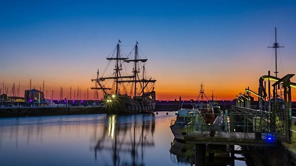 A harbor at sunset with a tall ship with multiple masts, smaller boats, and a reflecting calm water.