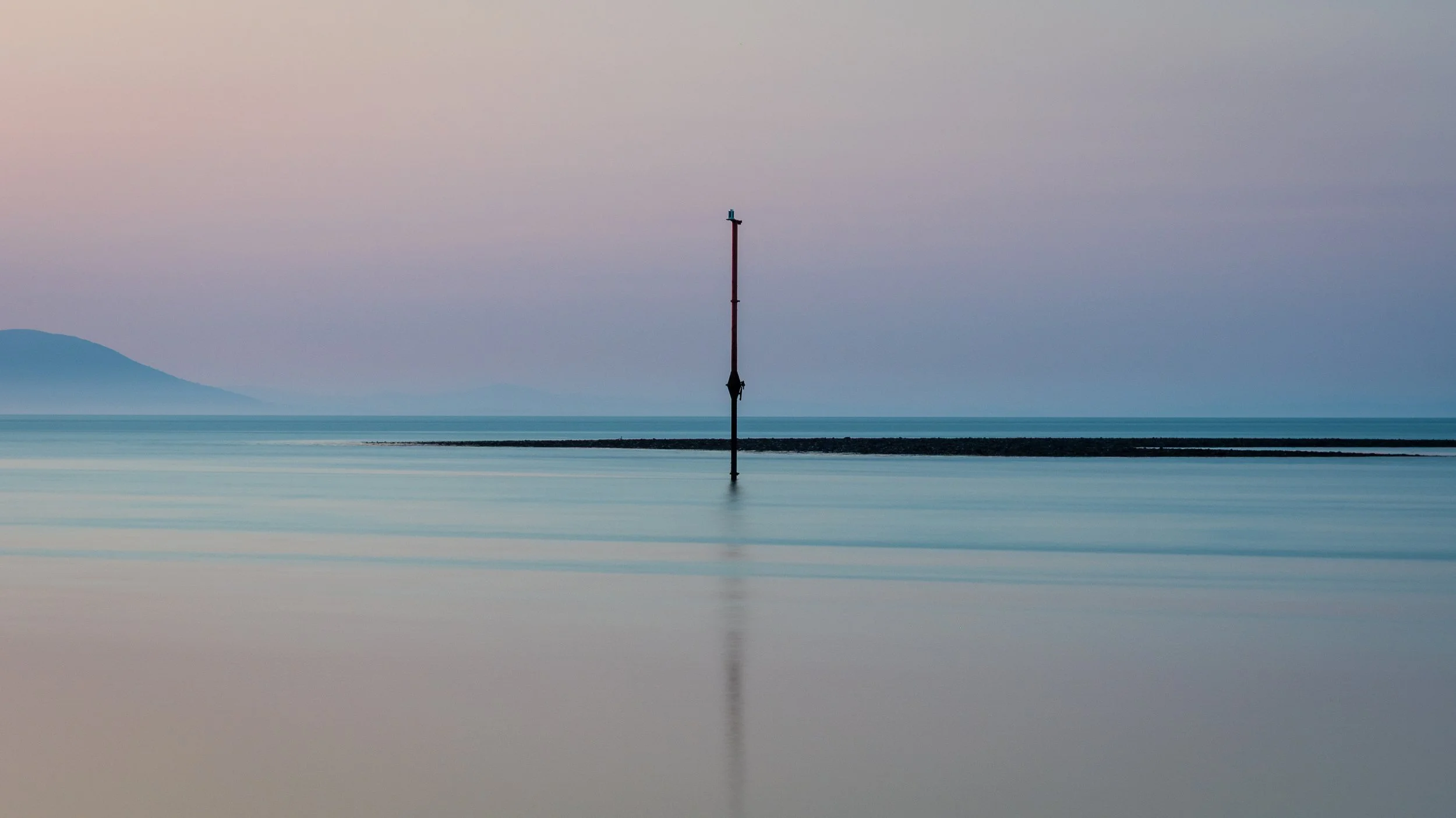 A calm body of water with a submerged navigation marker pole, with pastel sky and distant hills in the background.
