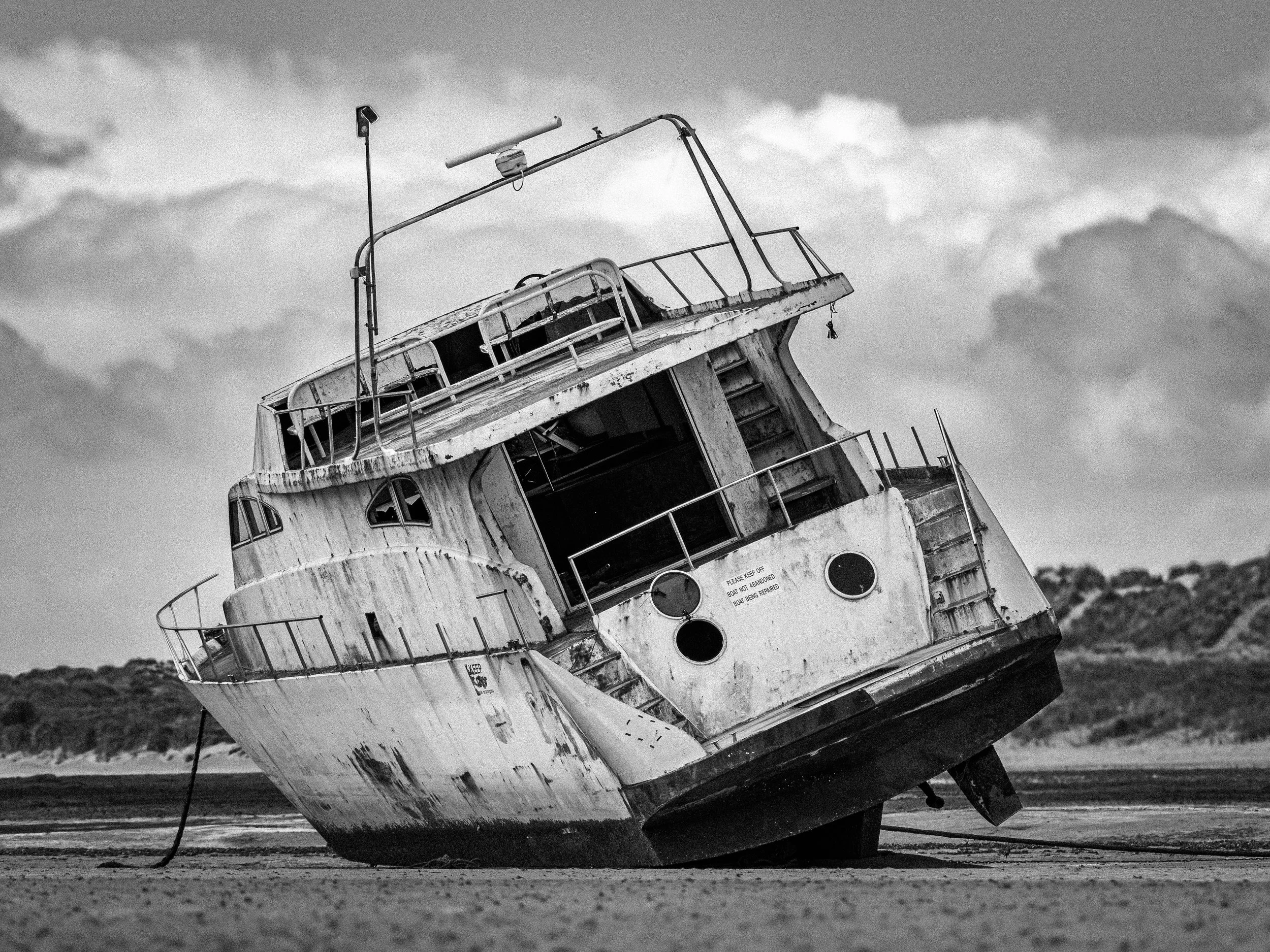 A large, abandoned boat tilted onto its side on a beach, appearing weathered and worn, with a cloudy sky in the background.