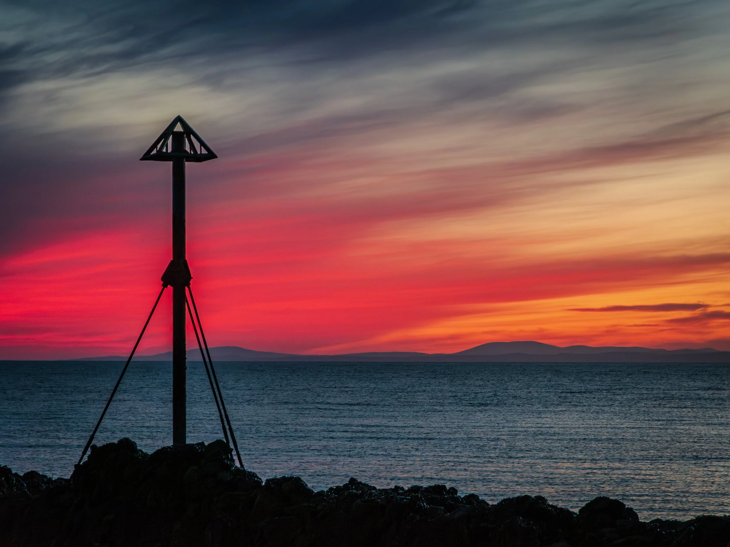 A navigation marker or buoy along the shoreline with a triangular top, silhouetted against a colorful sunset sky over the ocean with mountains in the distance.