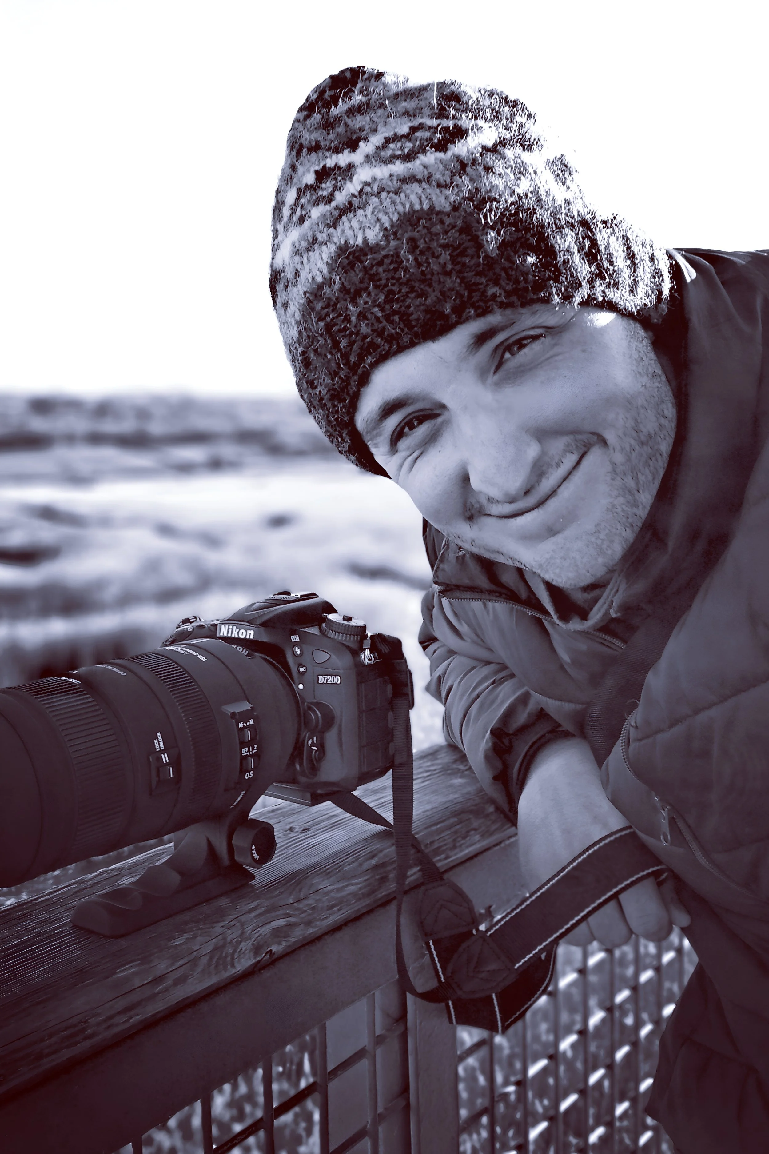A man with a knit hat and winter jacket smiling and looking at the camera while holding a camera with a large zoom lens, set outdoors in winter with snow in the background.