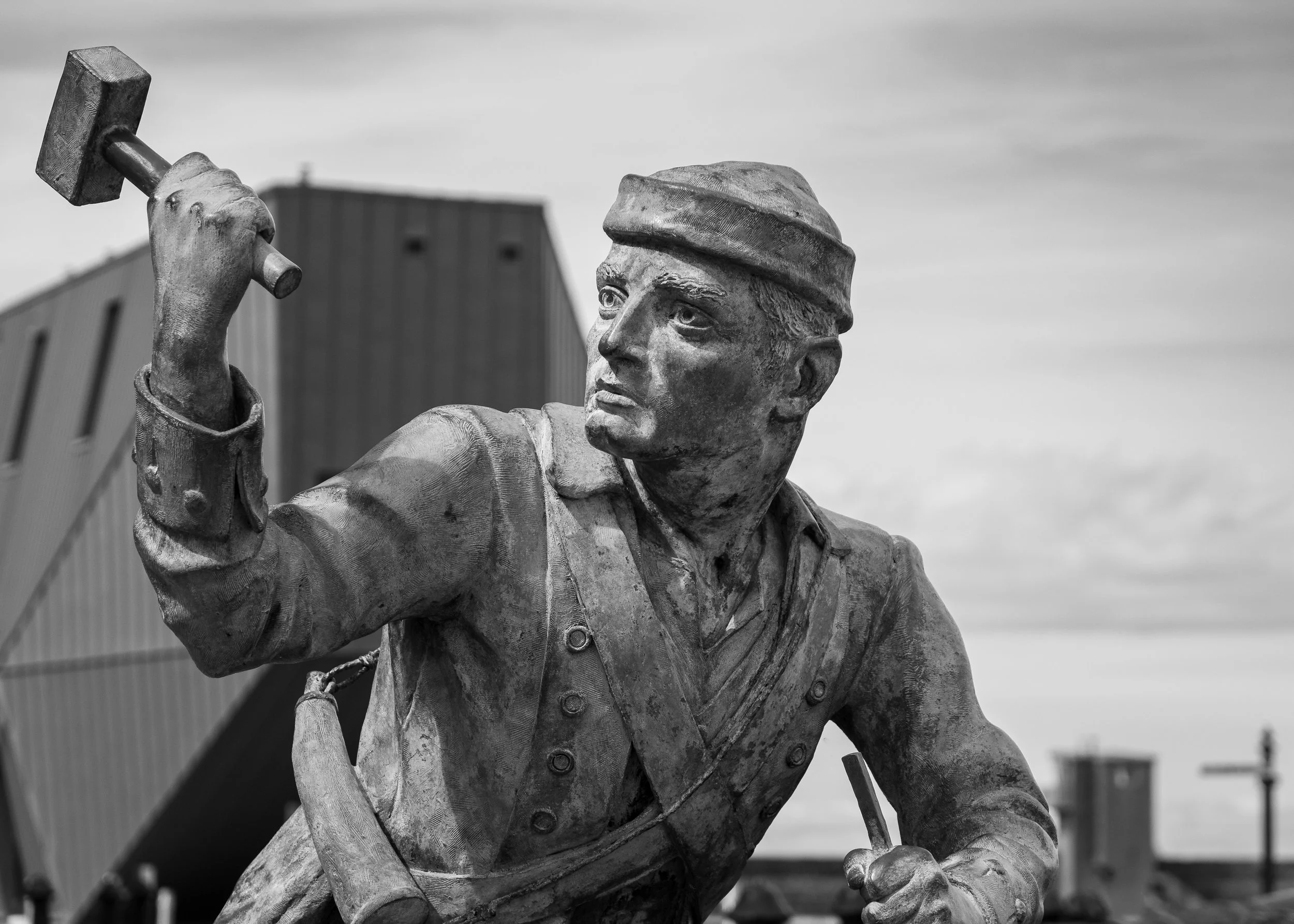 A black and white statue of a miner with a determined expression, wearing a cap, holding a hammer in his raised right hand, and gripping a tool in his left hand, set against an outdoor background. Location: Whitehaven Harbour, The Edge building.