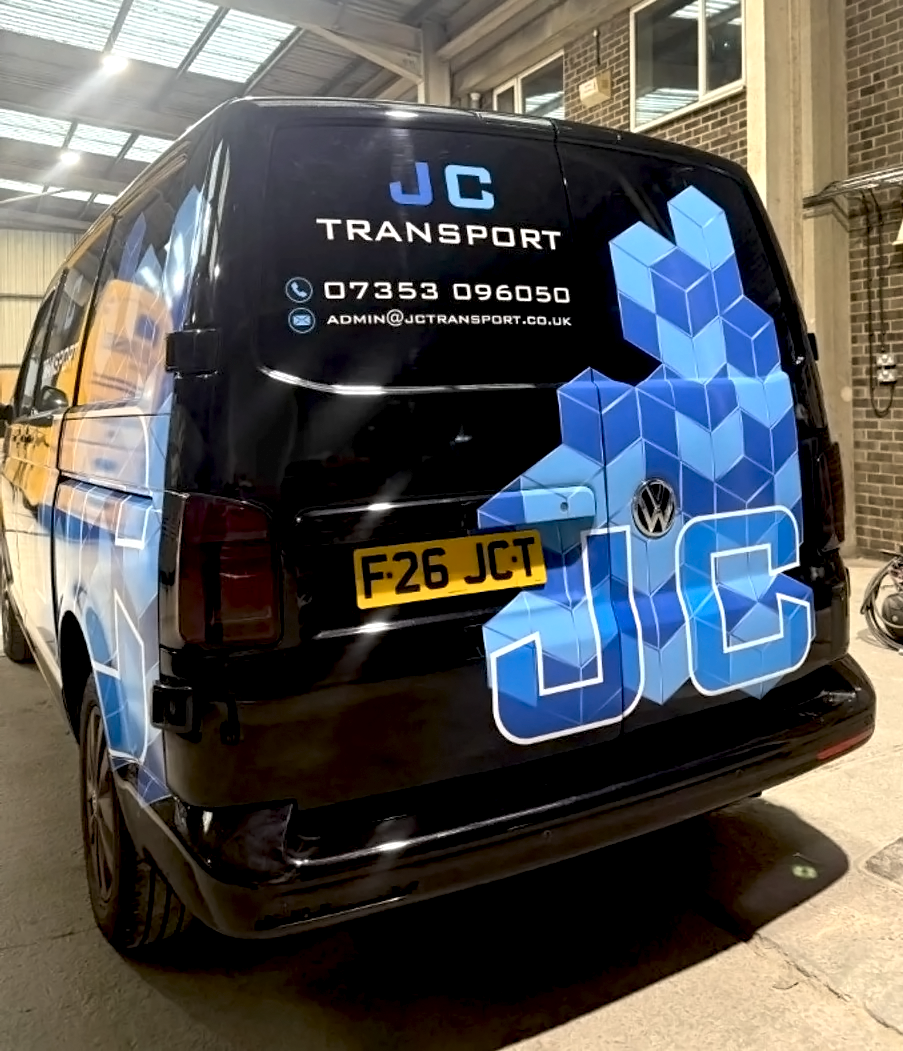 Black Volkswagen van with JC Transport logo and contact details, parked inside a garage with brick walls and a corrugated metal ceiling.