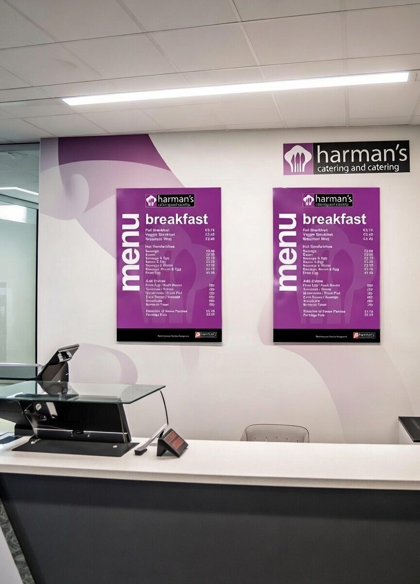 Interior of a food establishment with two purple menu boards displaying breakfast options and prices, a white counter with a cash register and a small digital device, and a white wall and ceiling with fluorescent lighting.