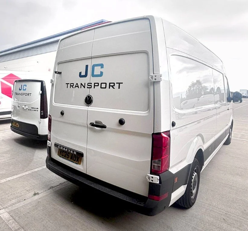 White transport van with JC Transport logo parked in a parking lot, with another JC Transport van nearby and a building with a pink stripe in the background.