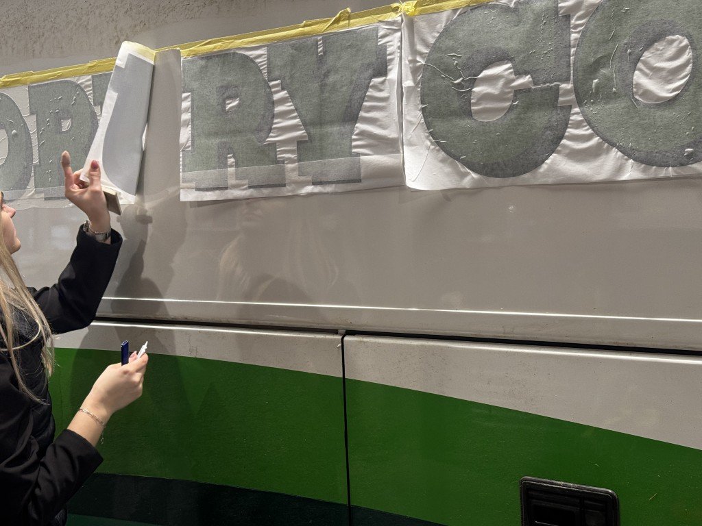 Person updating a large sign on the side of a bus or train, with the partial letters 'RY CG' visible.