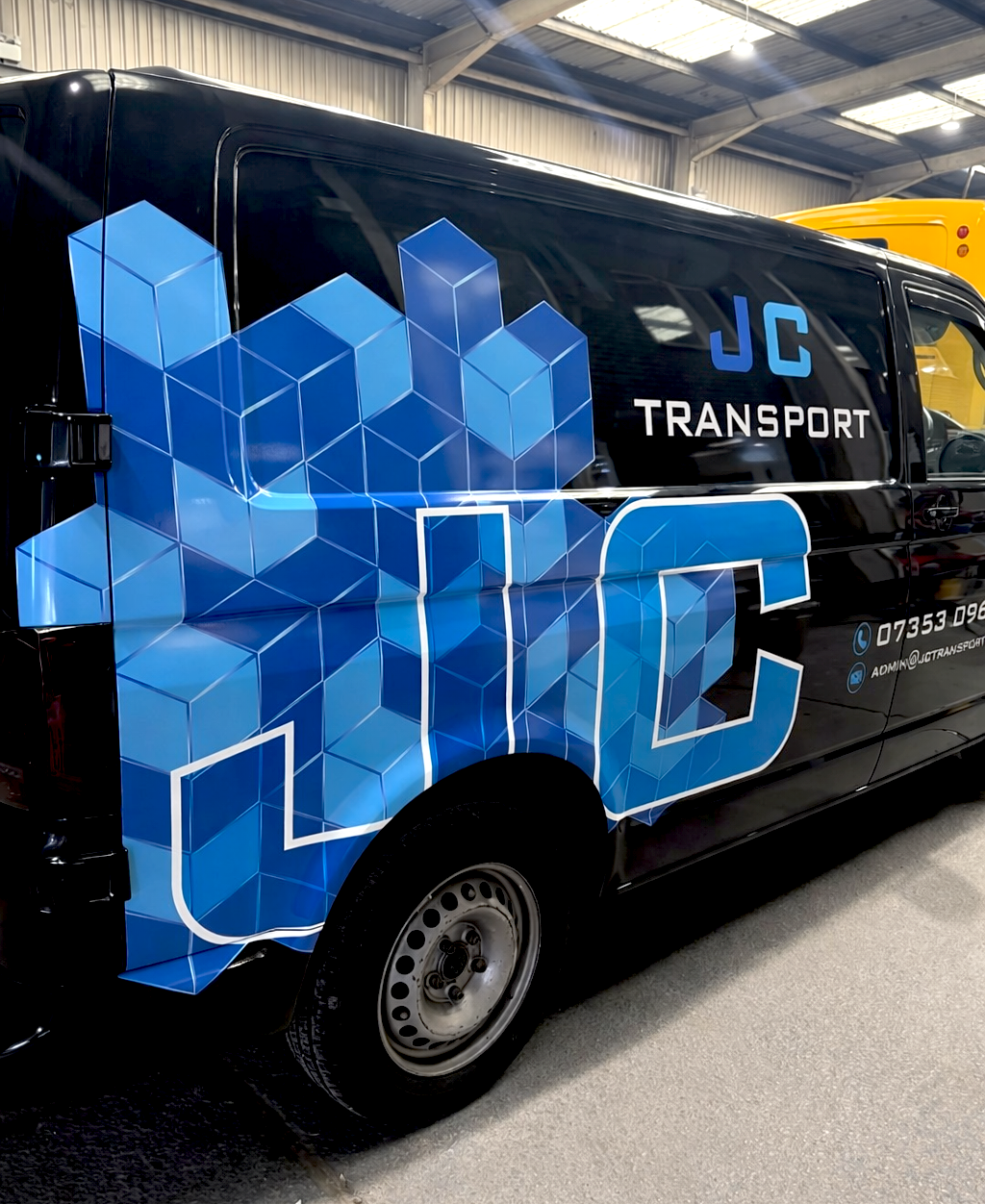 A black van with a blue geometric logo and the text 'JC Transport' on the side, parked inside a warehouse.