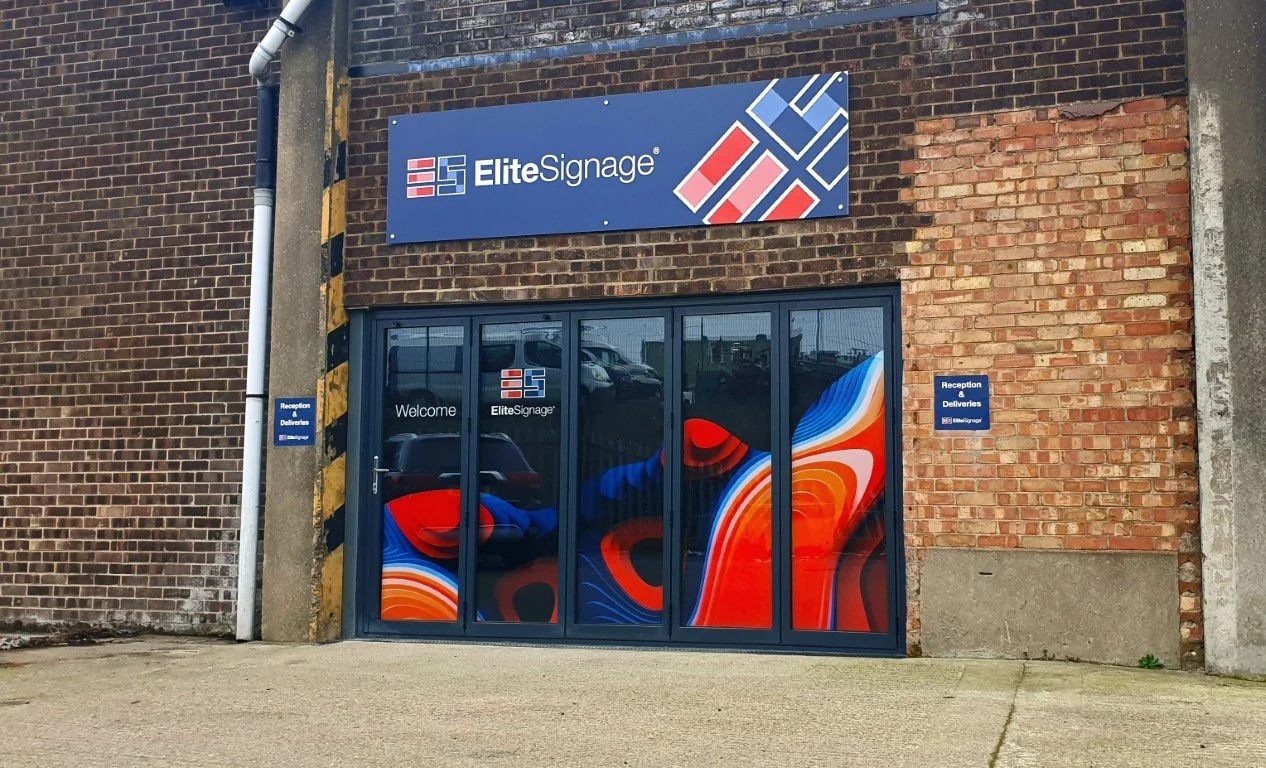 Exterior view of a storefront with a sign for Elite Signage, featuring a colorful abstract design on the glass doors and a blue sign on the brick wall indicating reception and deliveries.